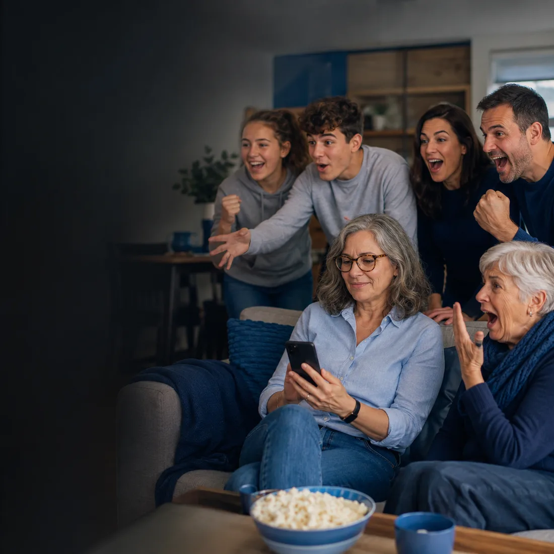 A family on the couch — three generations — one of them checking the tipping ladder on her phone while the others cheer.
